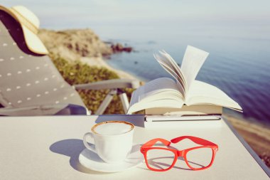 Tatillerde kitap okumak. Kahve fincanı ve kitaplar açık havada mavi deniz suyuna karşı. Kitap sayfaları rüzgarda çırpınıyor.