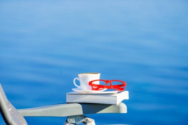 Tatilde okumak. Kahve fincanı, kitap ve kırmızı bardaklar mavi deniz suyuna karşı. Tatil gezisinde dinlenmek. Zihinsel sağlık sorunları.