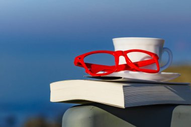 Tatilde okumak. Kahve fincanı, kitap ve kırmızı bardaklar mavi deniz suyuna karşı. Tatil gezisinde dinlenmek. Zihinsel sağlık sorunları.