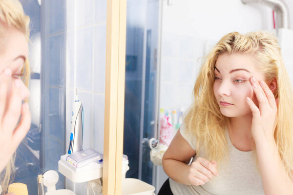 Sluggish woman looking at her skin condition in bathroom mirror. Sleepy languid female feeling tired, examing dark circles bags under eyes and wrinkles.