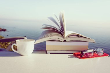 Tatillerde kitap okumak. Kahve fincanı ve kitaplar açık havada mavi deniz suyuna karşı. Kitap sayfaları rüzgarda çırpınıyor.