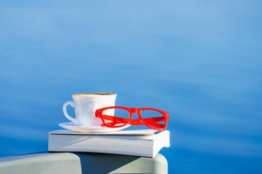 Tatilde okumak. Kahve fincanı, kitap ve kırmızı bardaklar mavi deniz suyuna karşı. Tatil gezisinde dinlenmek. Zihinsel sağlık sorunları.