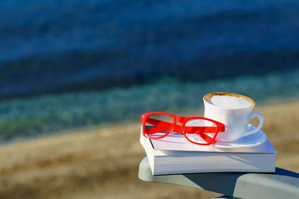 Spiritual healing, psyche silence. Coffee cup, book and red glasses against coast. Reading on vacation. Relaxation on holidays. Mental health break.