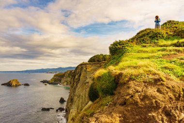 Kameralı kadın, İspanya 'nın kuzeyindeki Asturias kıyısındaki kayalık uçurumların deniz manzarasından fotoğraf çekiyor. Faro de Cabo Busto 'dan görüntü. Turist eğlencesi.