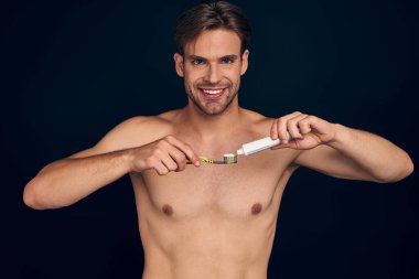 Handsome young bearded man isolated. Shirtless muscular man is standing on dark blue background with toothbrush and toothpaste in hand and smiling. Men care concept.