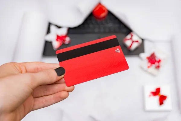 Female hand holding a red card for shopping in online stores, payment, spend money on a blurred notebook background with gift boxes on a white background.