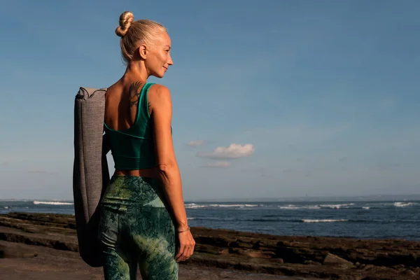 Güzel bir kız yoga minderiyle sahilde yürüyor. — Stok fotoğraf