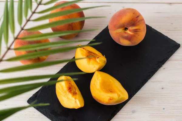 Three fresh ripe orange juicy peaches and their slices are lying on a black rectangular plate and a white wooden table. Top view through palm leaves. flat lay