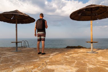 Hırvatistan 'ın deniz kıyısında hasır şemsiyeli ve bulutlu gökyüzü olan sırt çantalı genç beyaz bir adam. Arka görünüm