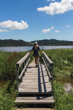 Young woman with hat walking on wooden bridge on educational trail Olsina from back. Sumava reserve, Czech summer landscape
