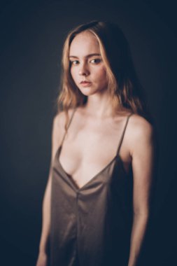 Avrupa Slav görünümlü ve kahverengi saçlı genç bir kız, bej rengi bir kimyevi elbise, kayışlı bir elbise, kameraya neşeyle bakıyor, oyunculuk yetenekleri ve yüzünde basit yüz ifadeleri, açık bir şekilde 