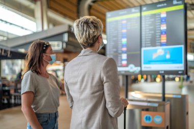 İki gezgin kadın koruyucu maskeler takıp Faro Havaalanı 'nda uçuş bilgi panosuyla tartışıyorlar.