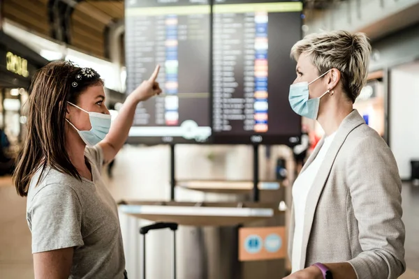 Two Travelling Women Wearing Protective Masks Discussing Flight Information Board — Stock Photo, Image