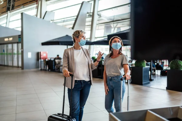 Two Travelling Women Wearing Protective Masks Discussing Flight Information Board — Stock Photo, Image