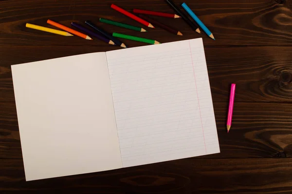 School notebook with oblique lines, colored pencils on the table wooden background. Blank sheet of paper in notebook on the desk. Back to school. School supplies. Top view. Copy space for text