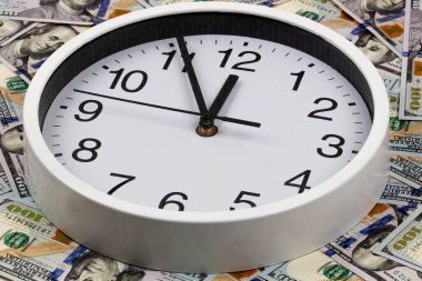 Vakit nakittir. Siyah sayılı büyük beyaz bir saat yüz dolarlık banknotlarla çevrilidir. İş ve finans kavramı. Saat 12 'ye 5 kala ABD doları arkaplanını gösteriyor.