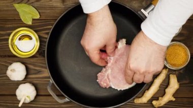 The cook puts raw meat steaks in a frying pan. Sliced pork meat in a cast iron skillet. Fry ingredients. Olive oil, bay leaf, garlic, mustard seeds, zira, ginger root, dry spices. Close-up. Top view