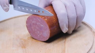 The cook cuts meat sausage into large pieces for a sandwich on a wooden table. A thick cured salami sausage made from fermented and dried meat. Pork, beef and veal salami on a kitchen board. Close-up