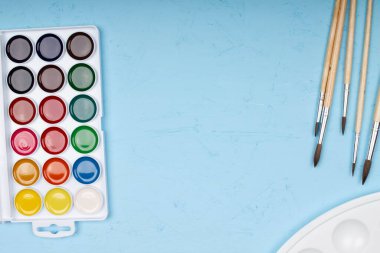 Okula dönelim. Fırçalar, boyalar ve paletler masanın üzerinde. Farklı boyutlarda fırçalar, güzel mavi bir arka plan çizmek için çok renkli parlak boyalar. Metin için boşluk. Üst görünüm