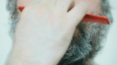 Adam sakalını tarıyor. Orta yaşlı bir adam büyük, dağınık gri kahverengi sakalını tarak ile tarıyor. Uzun bir sakalın kesilmesi, şekillendirilmesi ve berber dükkanında tımar edilmesi gerekiyor. Yakın plan.