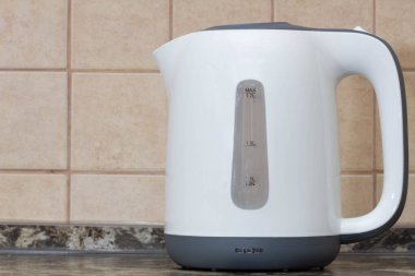 Electric kettle. White plastic electric kettle on the kitchen table against the background of beige tiles on the wall. Kitchen equipment. Boiling water for tea, coffee, hot drinks. Copy space for text