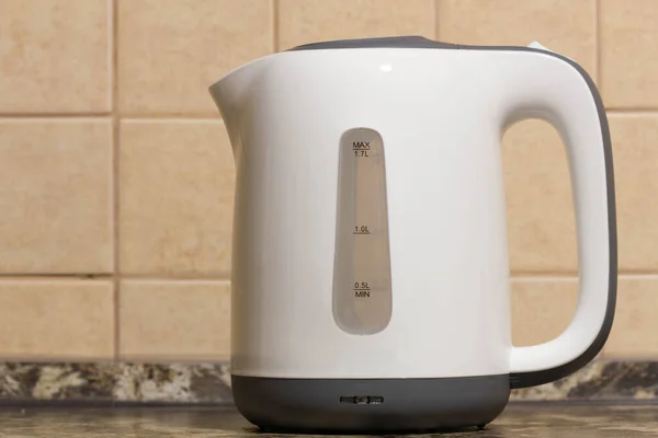 Electric kettle. White plastic electric kettle on the kitchen table against the background of beige tiles on the wall. Kitchen equipment. Boiling water for tea, coffee, hot drinks. Copy space for text