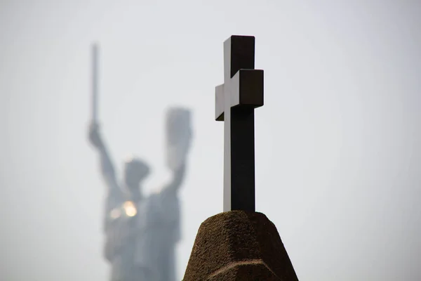 Cross in the cemetery. A Catholic cross on the grave against the background of a military monument to the Motherland. Christian grave cross in the bright sun. Black stone cross against the blue sky