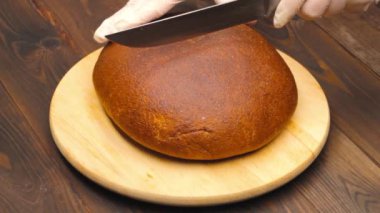 Slicing bread. Hands in white gloves cut a loaf of brown bread with a large kitchen knife on a cutting board against the background of a brown wooden table. Healthy food. Cooking food in the kitchen