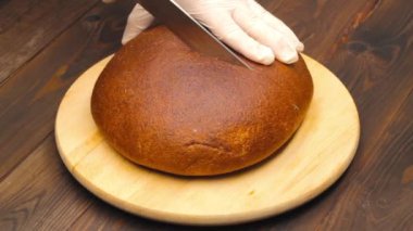 Slicing bread. Hands in white gloves cut a loaf of brown bread with a large kitchen knife on a cutting board against the background of a brown wooden table. Healthy food. Cooking food in the kitchen