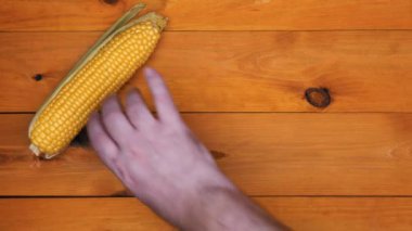 Hands put corn and knife on the table. A man lays out corn on the cob with green leaves and husks and a large knife on the table on an orange wooden background. Fresh corn for cooking. Health food