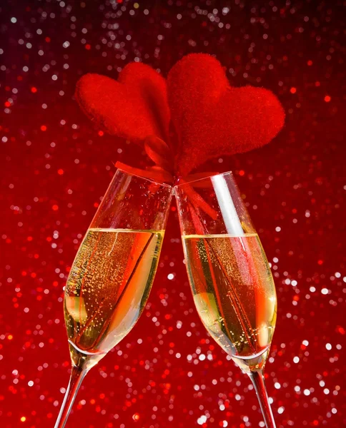 Two champagne flutes with golden bubbles and red velvet hearts make cheers on red bokeh background — Stock Photo, Image