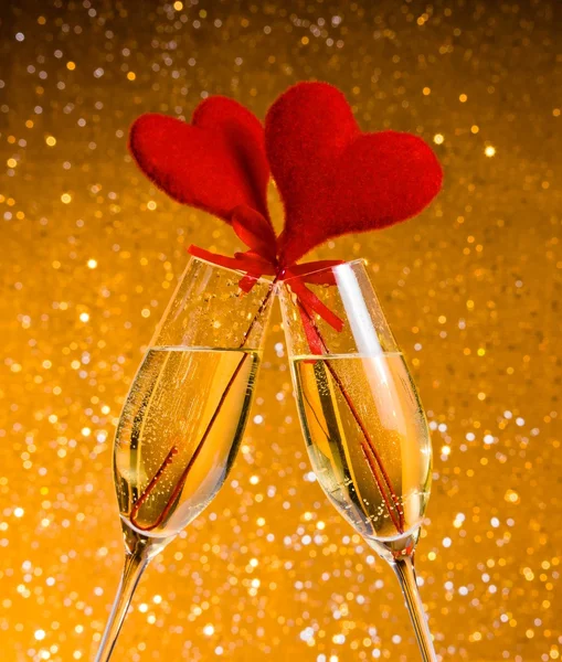 Two champagne flutes with golden bubbles and red velvet hearts make cheers on golden bokeh background — Stock Photo, Image