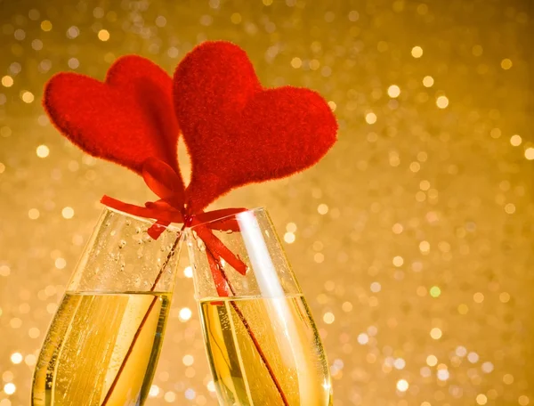 Two champagne flutes with golden bubbles and red velvet hearts make cheers on golden bokeh background — Stock Photo, Image