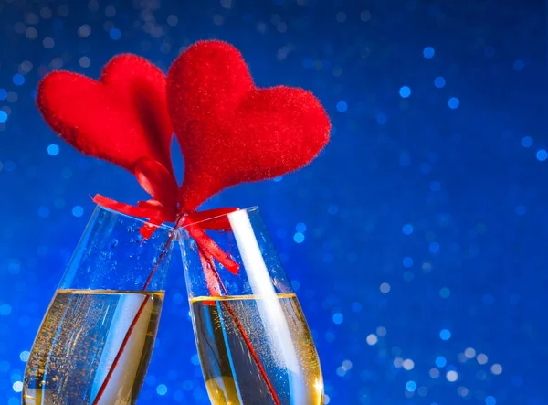 Two champagne flutes with golden bubbles and red velvet hearts make cheers on blue bokeh background — Stock Photo, Image