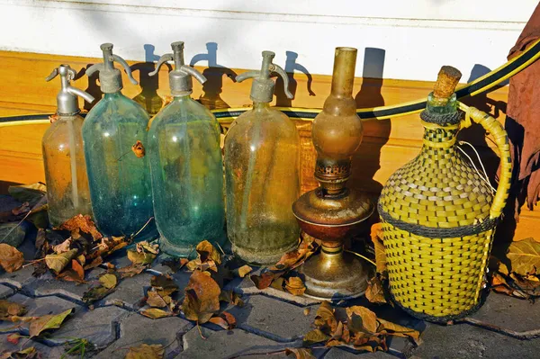 Four old glass siphons, a kerosene lamp and a glass bottle for wine in a plastic braid among the dry fallen leaves on the pavement