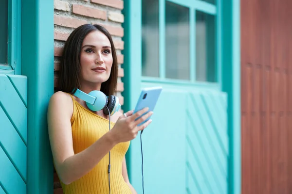 Гарненька молода жінка з смартфоном і навушниками, спираючись на стіну — стокове фото