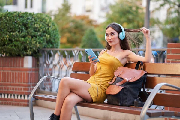 Красивая молодая женщина сидит на скамейке в городе и слушает музыку в наушниках. — стоковое фото