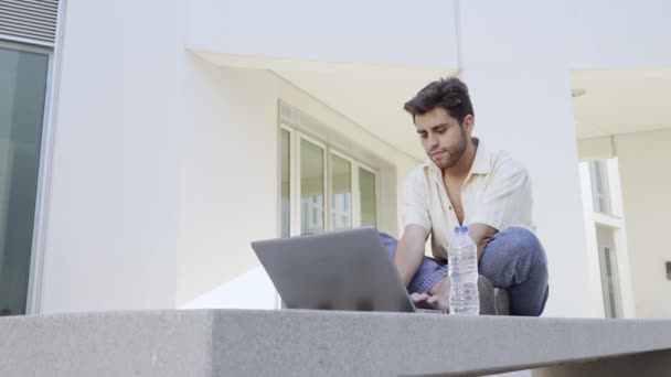 Молодой бородатый мужчина печатает на своем ноутбуке, сидя на скамейке — стоковое видео