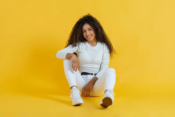 Portrait of young woman sitting on studio floor smiling and looking at camera with yellow background — Foto de Stock