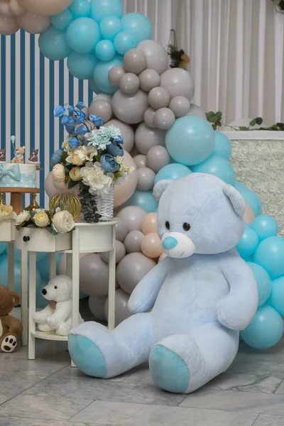 Teddy bear sits on background with different colors of balloons, birthday garland, blue and white flowers. Celebrating first year baby\'s life. Decorated photo zone