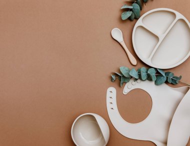 Kahverengi arka planda bebek maması aksesuarı. Nötr silikon önlük ve kase. Çocuk bulaşık takımı, bebek maması. 