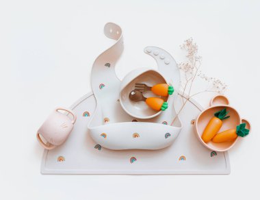 Bej arka planda bebek önlükleri ve plakası olan düz bir kompozisyon. Yemek servisi. Çocuk menüsü, beslenme ve beslenme kavramı