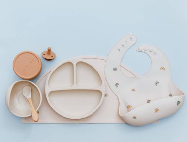 Mavi arka planda silikon bebek önlüğü ve bej bulaşık takımı olan düz bir kompozisyon. Düz yatış, üst görünüm