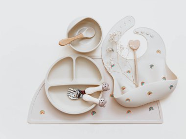 Bej arka planda bebek önlükleri ve plakası olan düz bir kompozisyon. Yemek servisi. Çocuk menüsü, beslenme ve beslenme kavramı