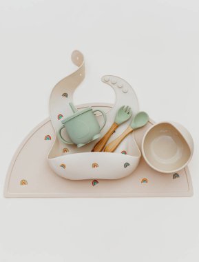 Bebek için silikon masa takımı, önlükler ve bej arka planda plaka içeren düz bir kompozisyon. Yemek servisi. Çocuk menüsü, beslenme ve beslenme kavramı