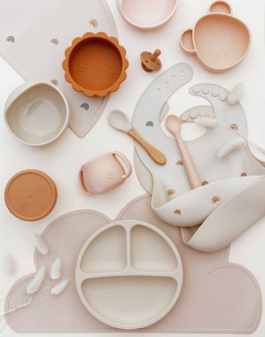 Üst görünüm silikon tabak ve kaşık. Bebeğe hizmet etmek, ilk beslenme konsepti. Masa örtüsü ve önlük açık arka planda. Düz konum, boşluk kopyala, üst görünüm