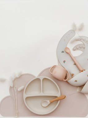 Top view modern silicone dishware plate and spoon. Serving baby, first feeding concept. Kids tableware and bib on light background. Instagram use. Flat lay, copy space, top view