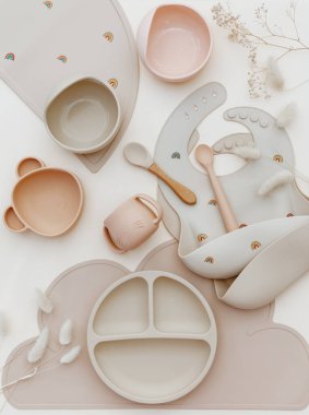 Üst görünüm silikon tabak ve kaşık. Bebeğe hizmet etmek, ilk beslenme konsepti. Masa örtüsü ve önlük açık arka planda. Düz konum, boşluk kopyala, üst görünüm