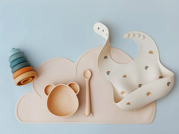 Pastel silicone collection of tableware, bibs, accessories, Montessori toys for children on blue background. Baby accessories, tableware concept.