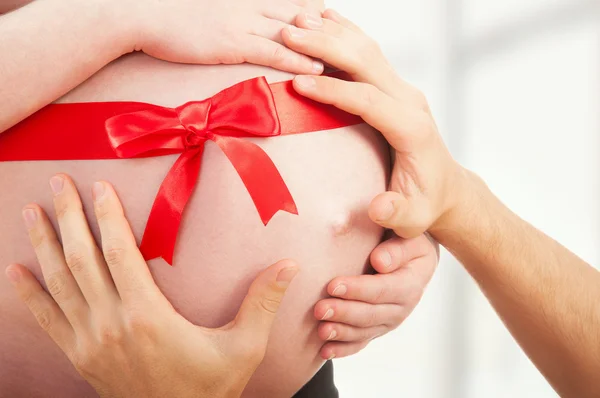 Беременный живот с красной лентой и руками родителей — стоковое фото